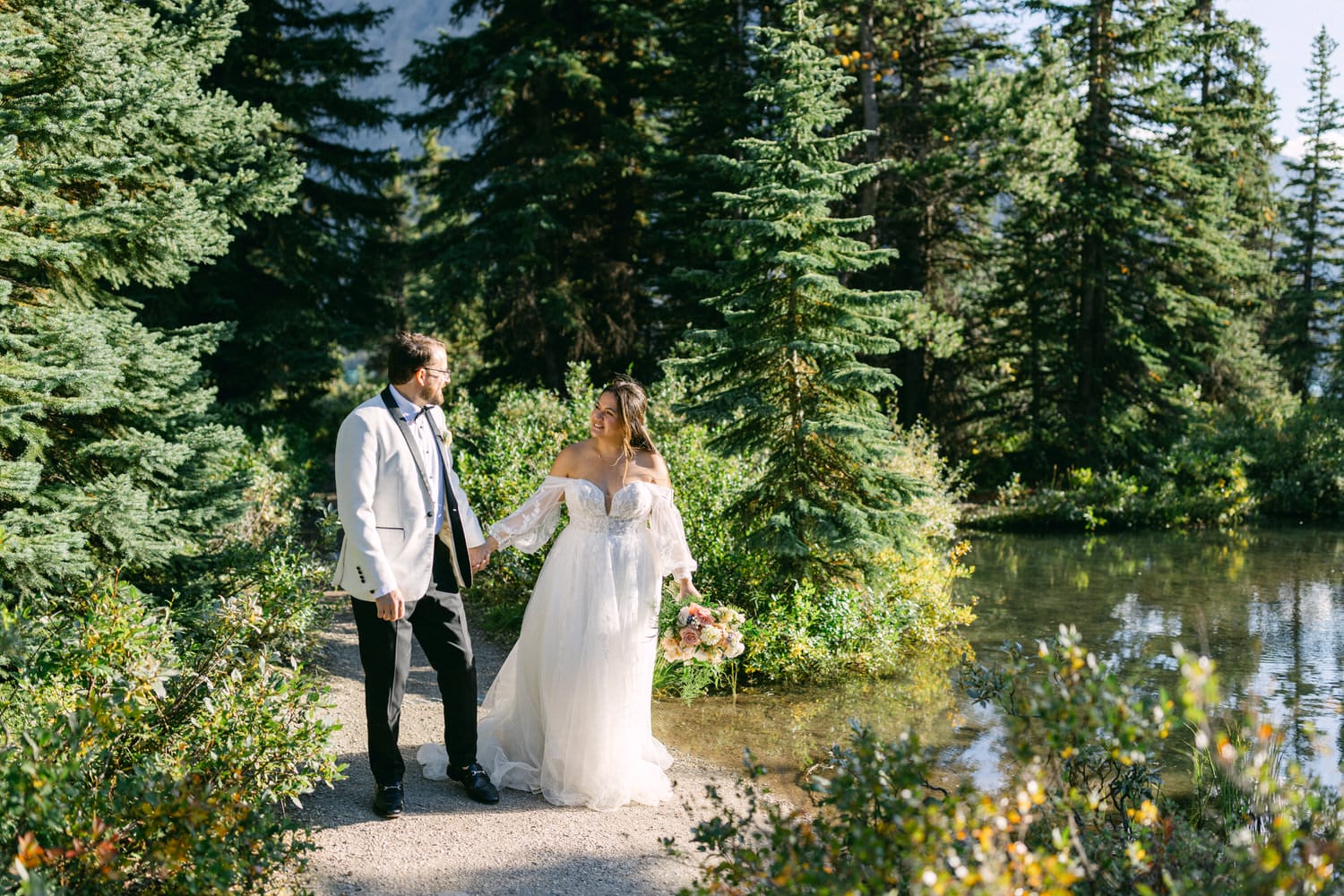 A bride and groom holding hands by a serene lake surrounded by lush greenery