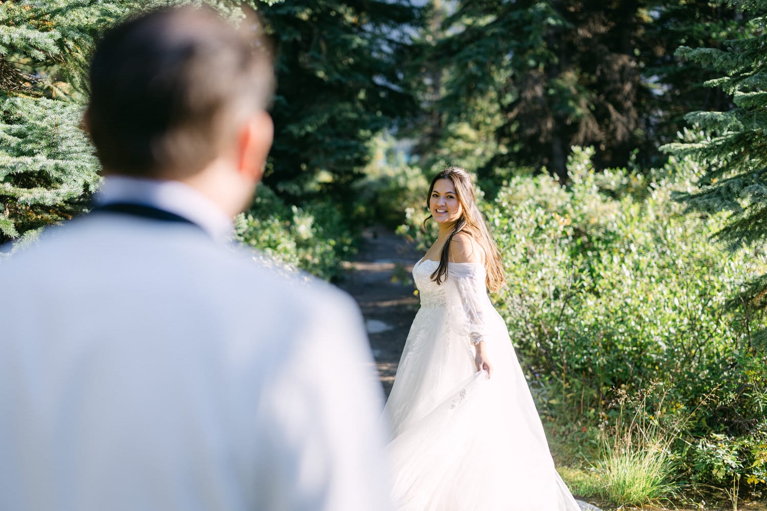 A bride in a white gown smiling over her shoulder at the groom whose back is to the camera, amidst lush greenery.