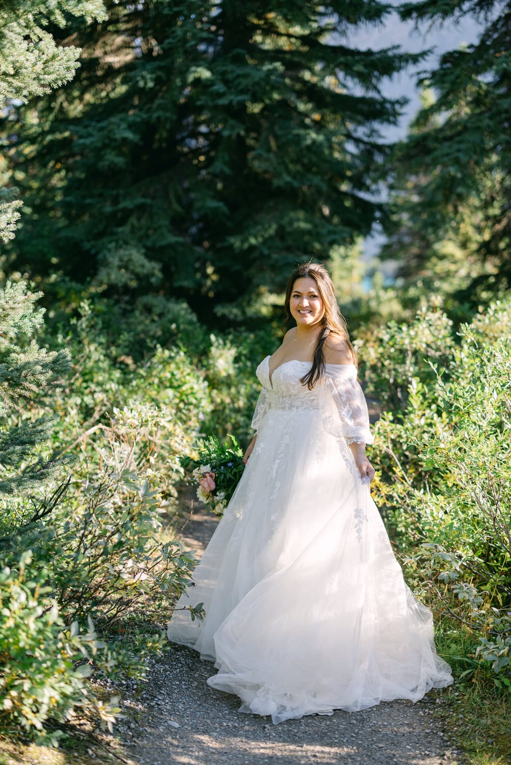 A smiling bride in a white dress standing on a forest pathway surrounded by greenery.