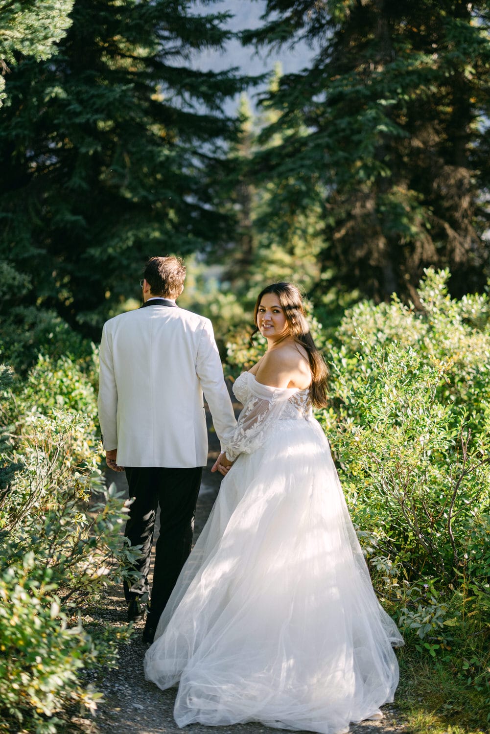 Bride in a flowing gown holding hands with groom, walking down a forested path.