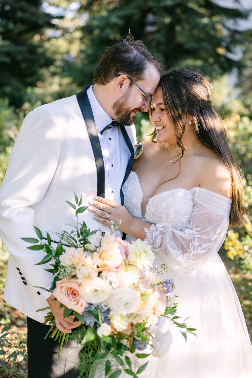 A bride and groom affectionately touching foreheads, holding a bouquet, with the bride in an off-shoulder lace gown and the groom in a white tuxedo with a black bow tie.