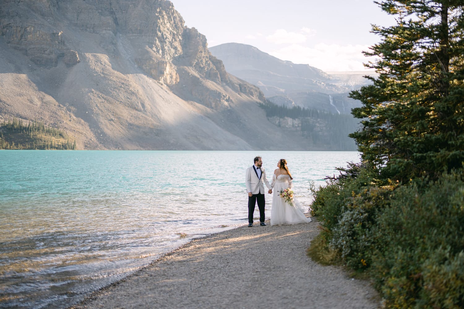 A bride and groom holding hands by a crystal blue lake with mountains in the background.