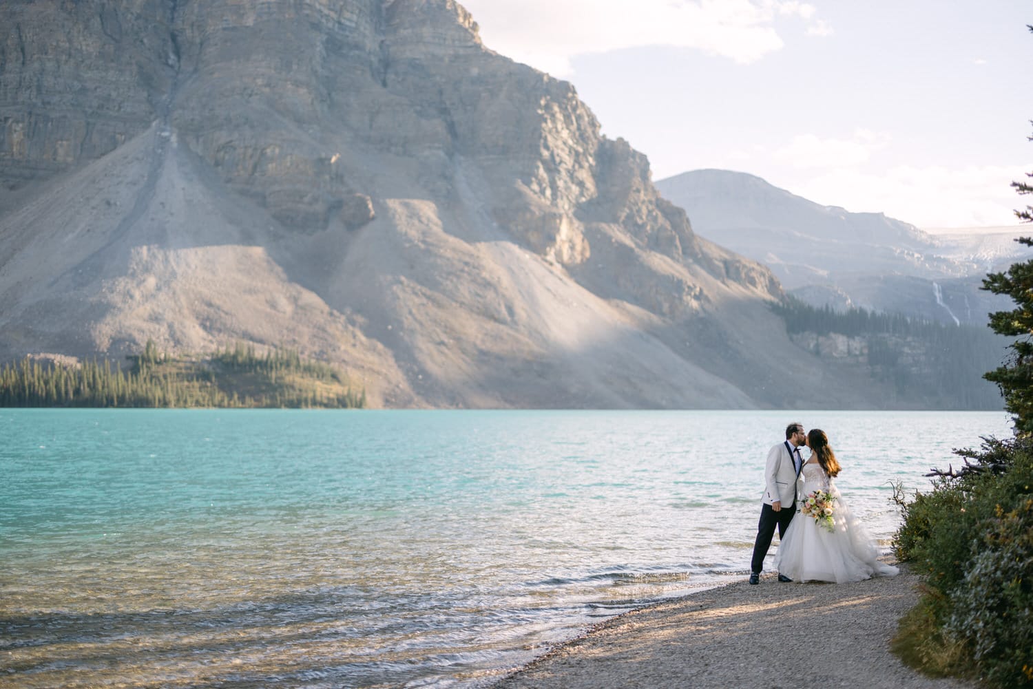 A couple in wedding attire sharing a kiss by a serene mountain lake with towering peaks in the background.