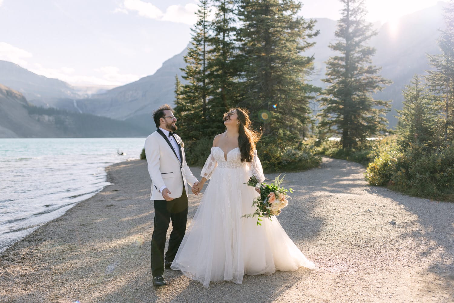 A bride and groom holding hands and smiling at each other on a picturesque lakeside path surrounded by mountains and trees.