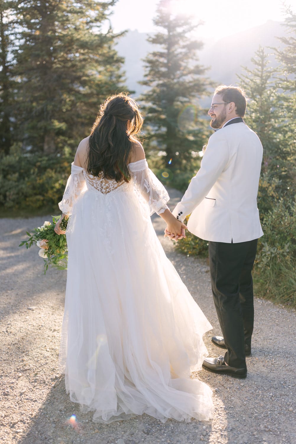 A bride and groom holding hands and walking together on a pathway surrounded by nature, with the warm glow of sunset in the background.