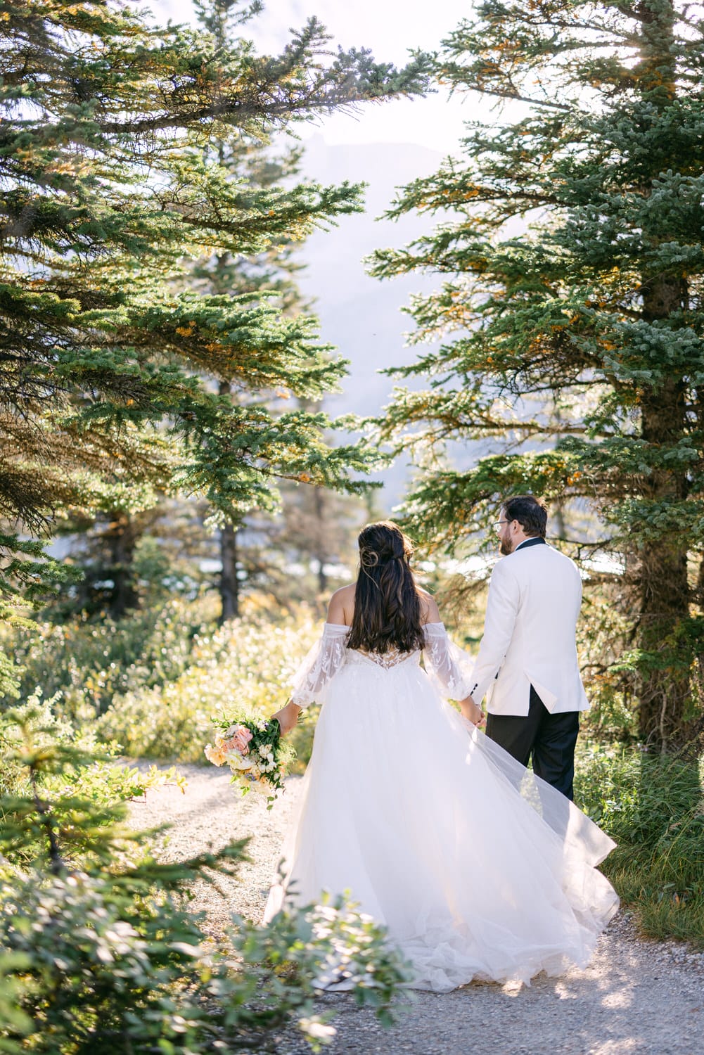 A bride and groom holding hands, walking down a forest path surrounded by trees, with the bride carrying a bouquet.