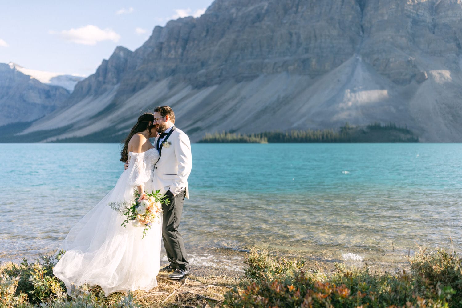 A bride and groom embracing by a turquoise lake with towering mountains in the background