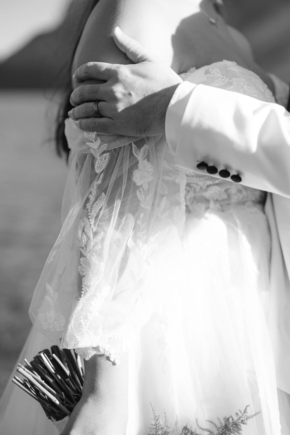 Black and white close-up of a bride and groom embracing on their wedding day, focusing on their hands and the details of the wedding attire.