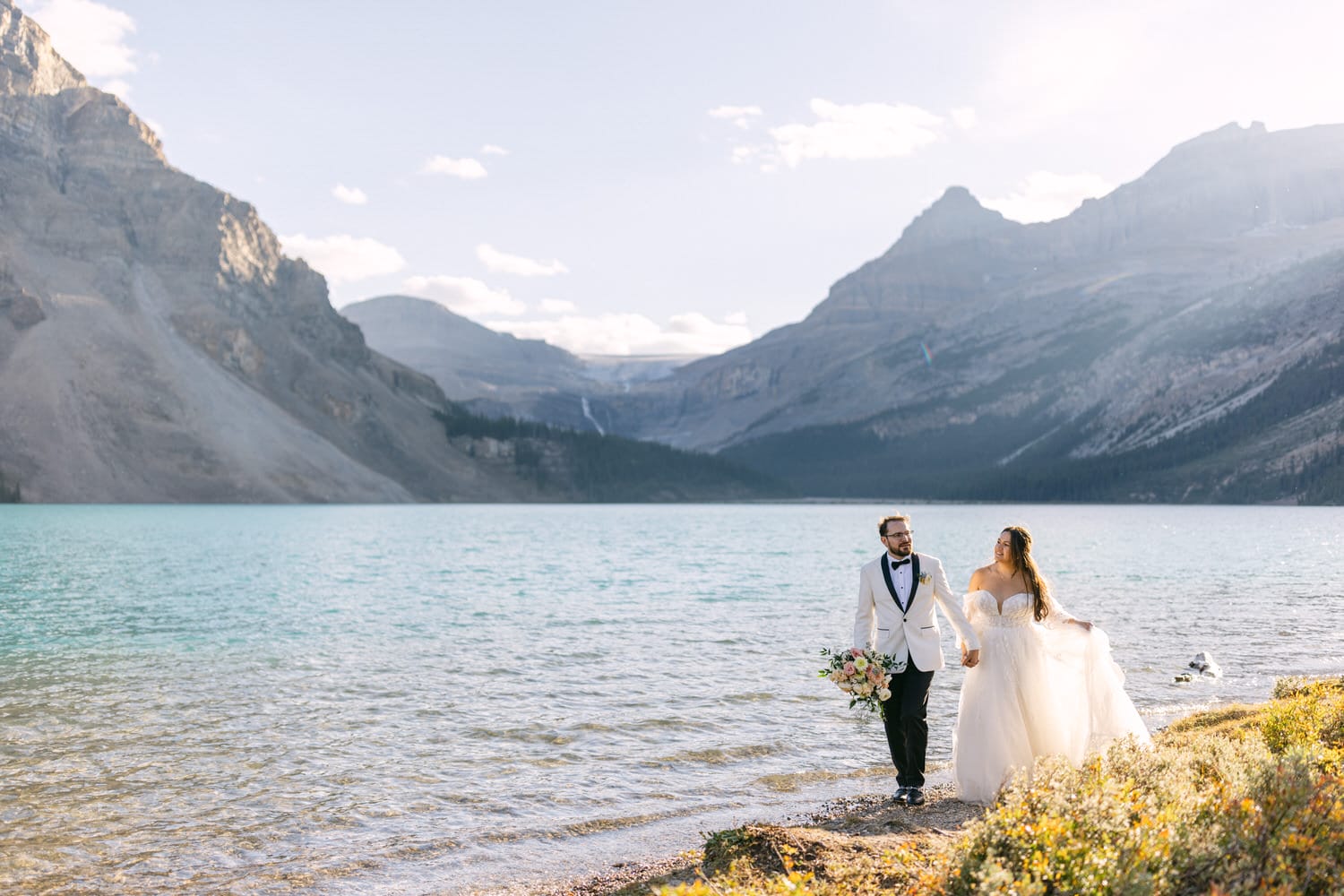 A bride and groom holding hands by an alpine lake with mountains in the background