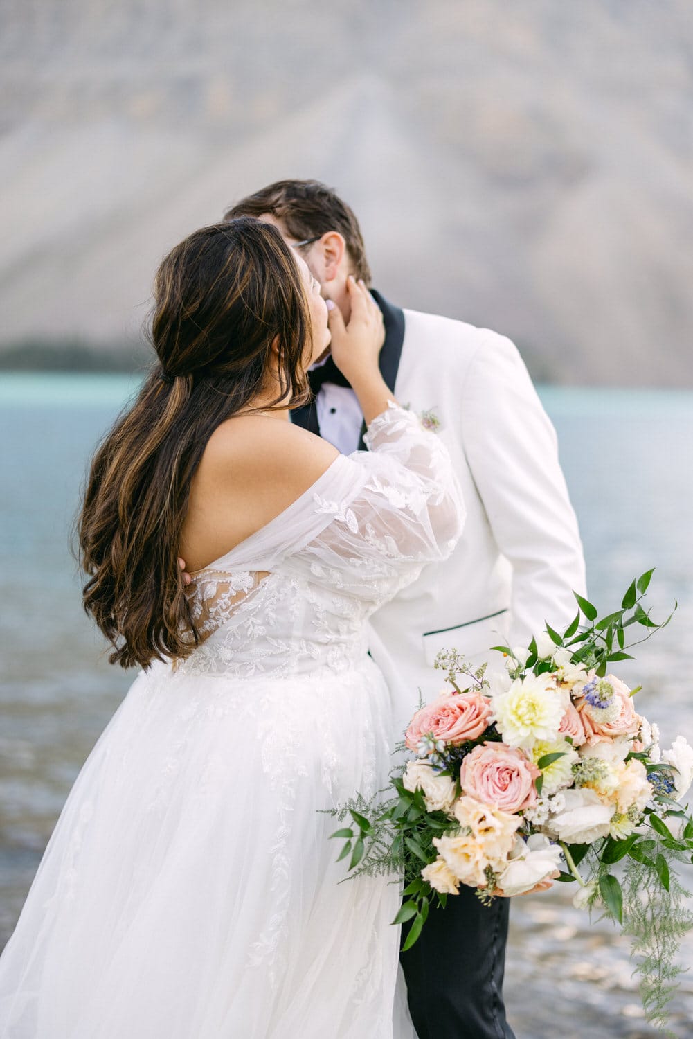 Bride in a white gown kissing the groom in a white jacket by a mountain lake, holding a bouquet of flowers.