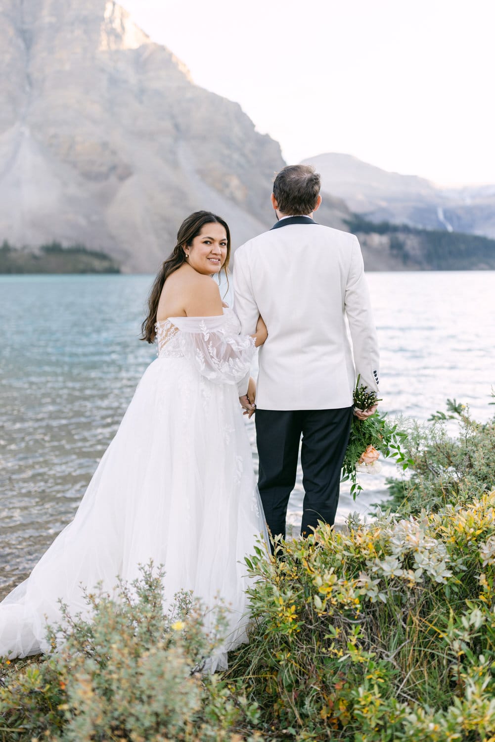 Bride in a white off-shoulder gown smiling at the camera while holding hands with a person in a white suit jacket, standing by a lake with mountains in the background.