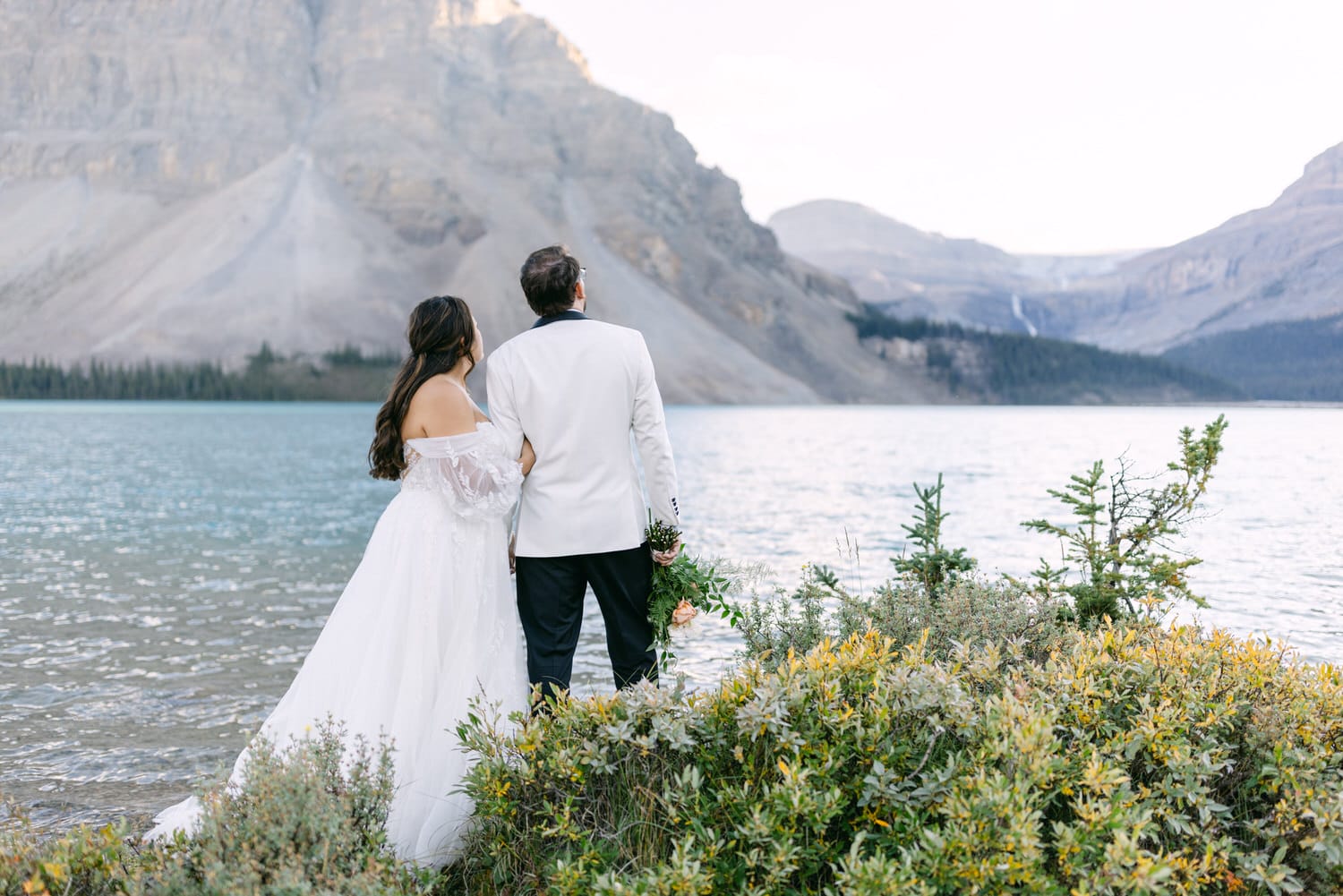 A bride in a white flowing dress and a groom in a white jacket stand together facing a tranquil mountain lake with lush greenery in the foreground.