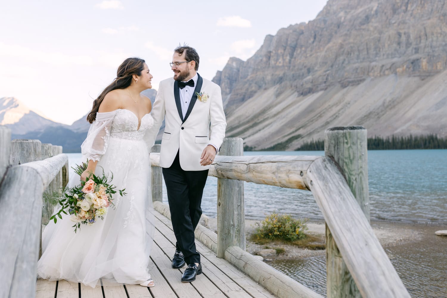 Bride and groom walking hand in hand on a wooden jetty by a mountain lake.