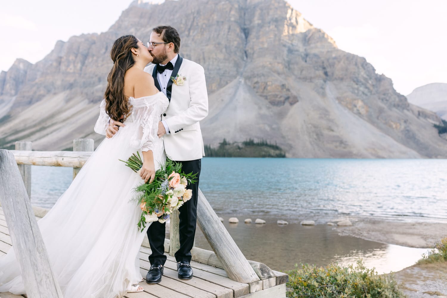 A bride and groom share a kiss on a wooden dock with a scenic mountain lake backdrop.