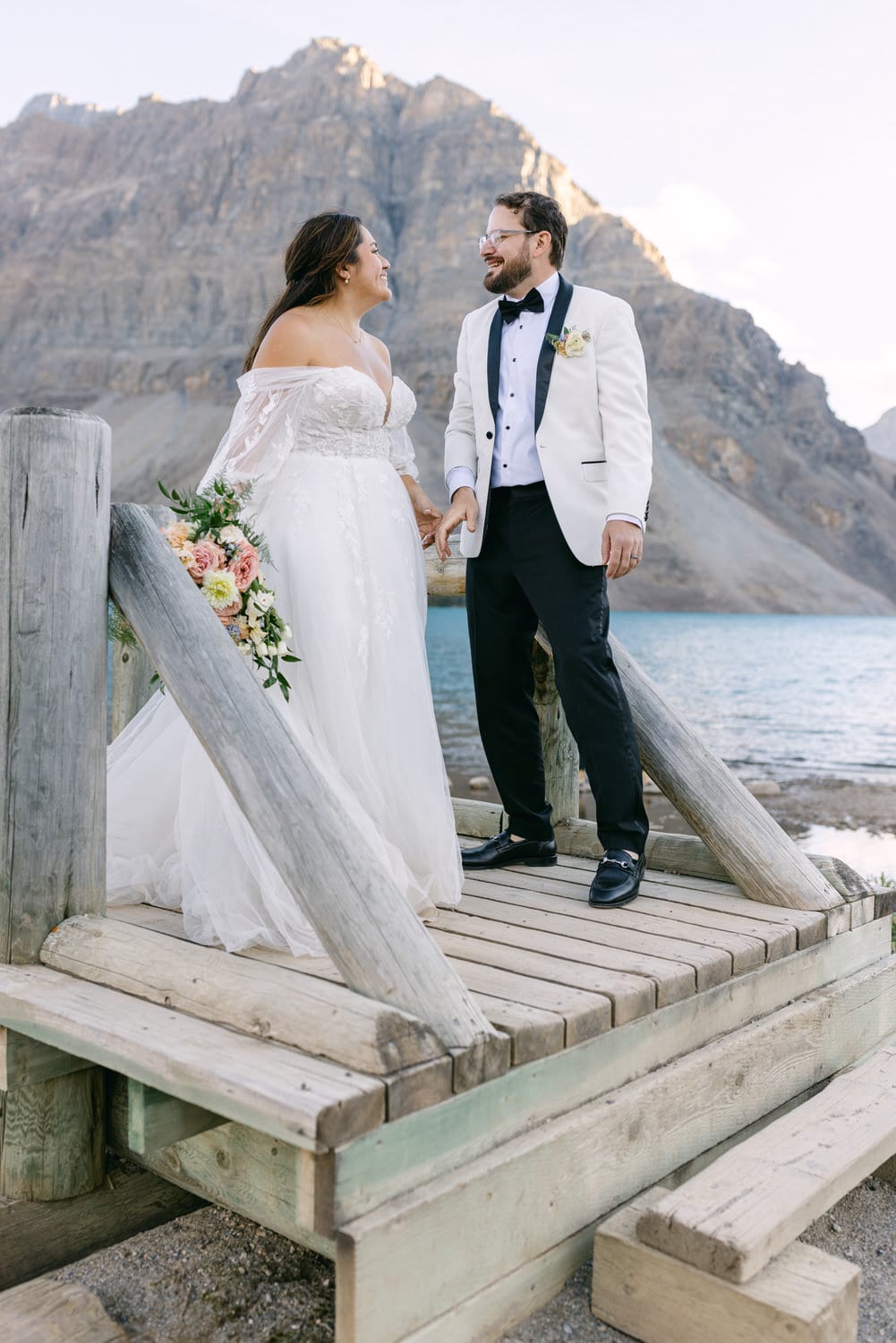 A happy bride and groom holding hands on a wooden pier by a mountain lake.