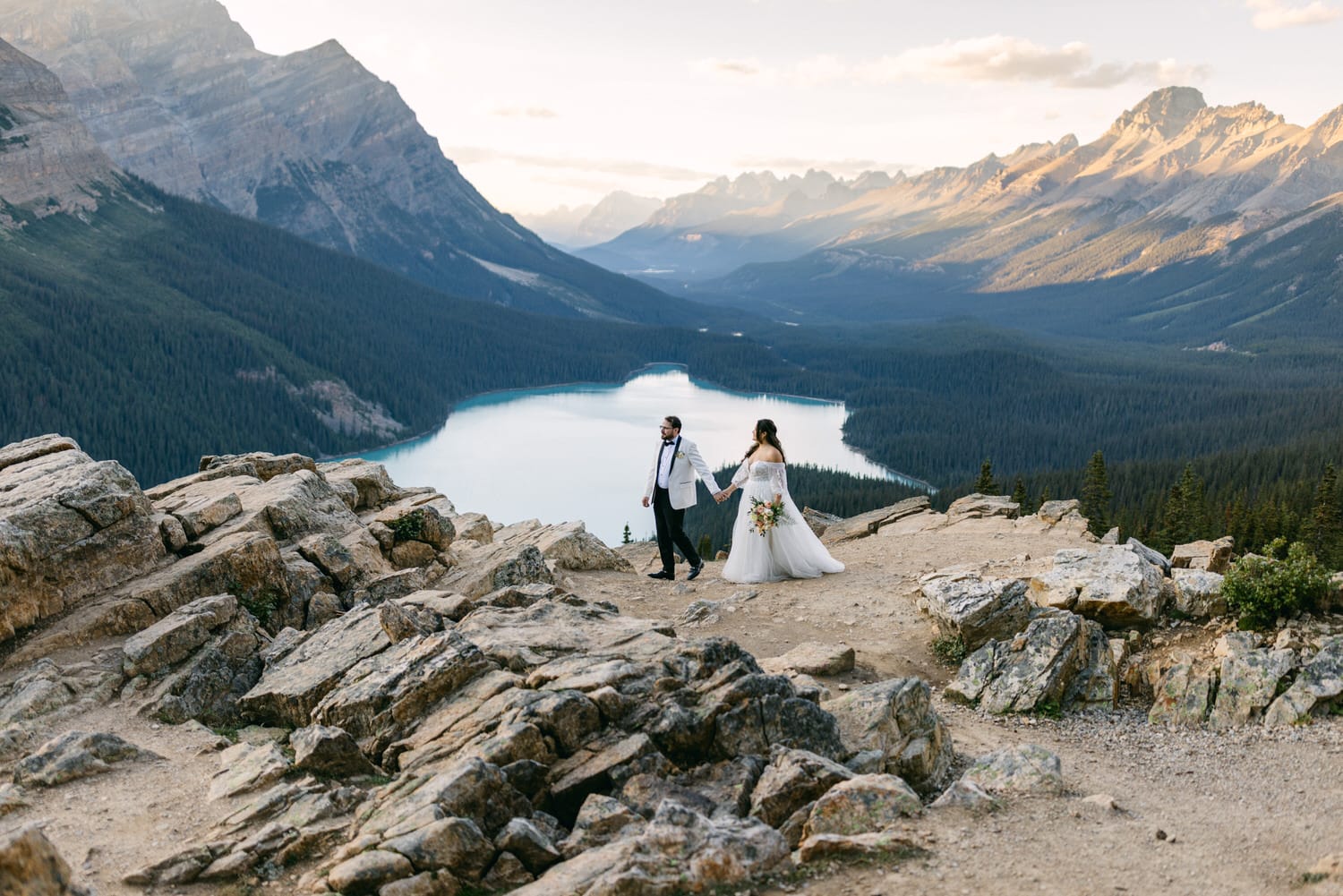 A bride and groom holding hands on a mountain overlook with a scenic lake and mountain range in the background at sunset.