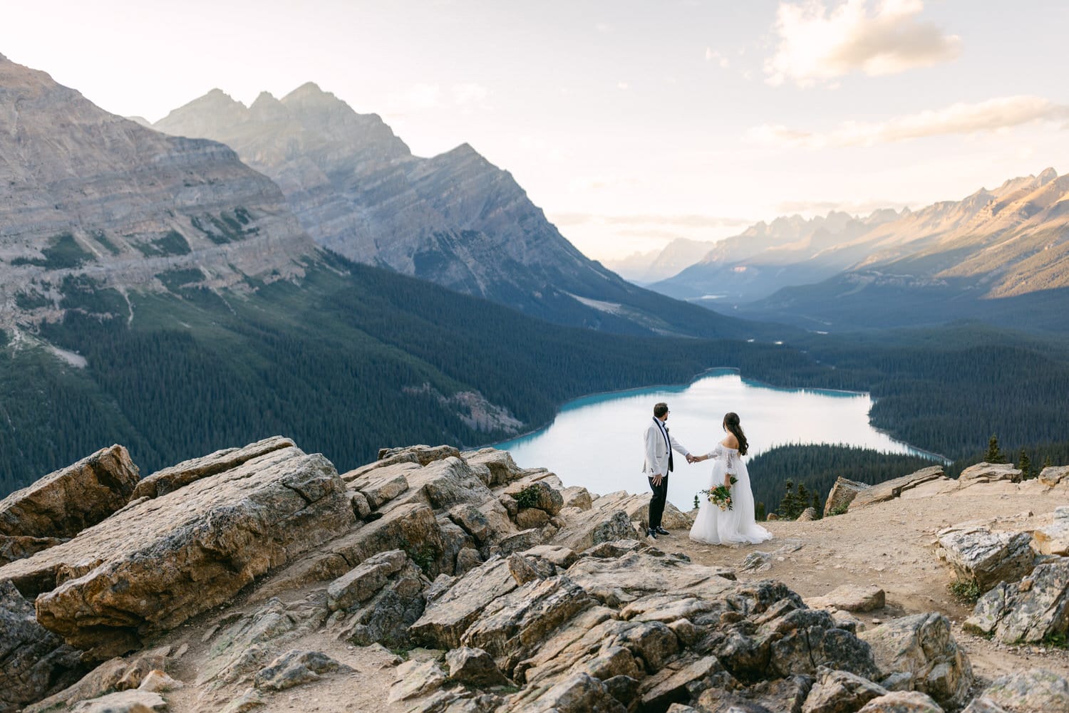 Bride and groom holding hands on a mountain overlook with a panoramic view of a lake and mountain range at dusk