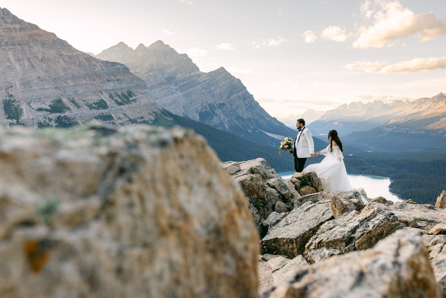 A bride and groom holding hands on a mountain summit with majestic views of rocky peaks and a serene lake in the background at sunset.