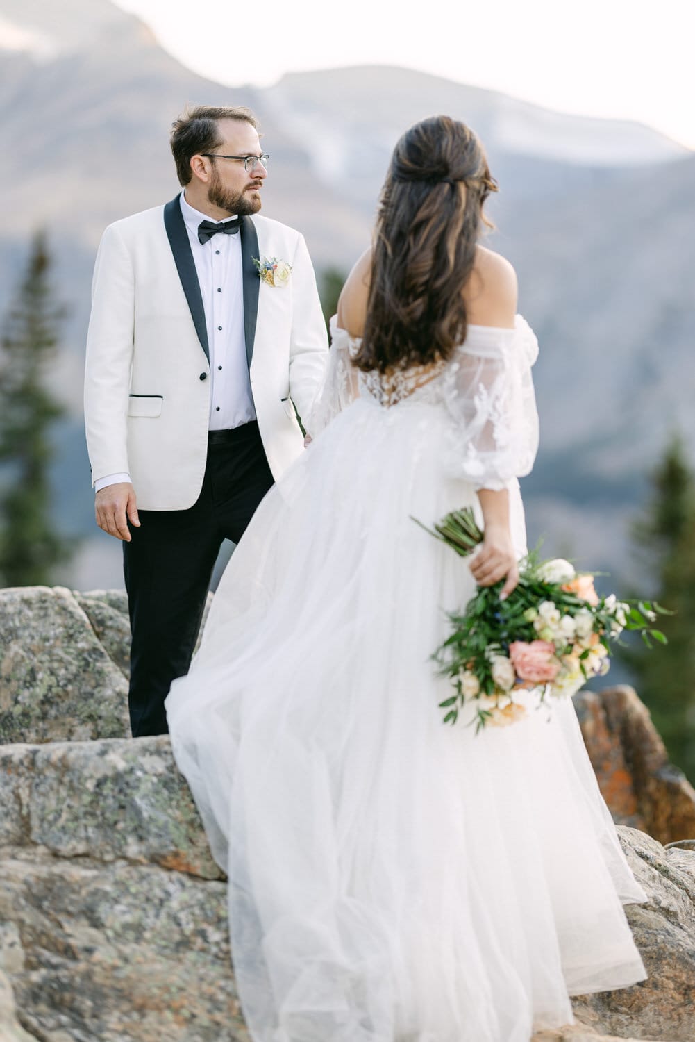 A groom in a white tuxedo jacket and bride in a flowing white gown with a bouquet, standing on rocky terrain with mountain scenery in the background.