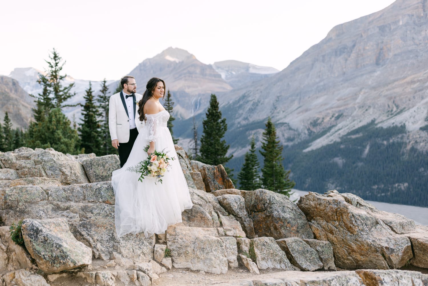 A bride and groom standing on rocky terrain with a mountainous backdrop, the bride holding a bouquet.