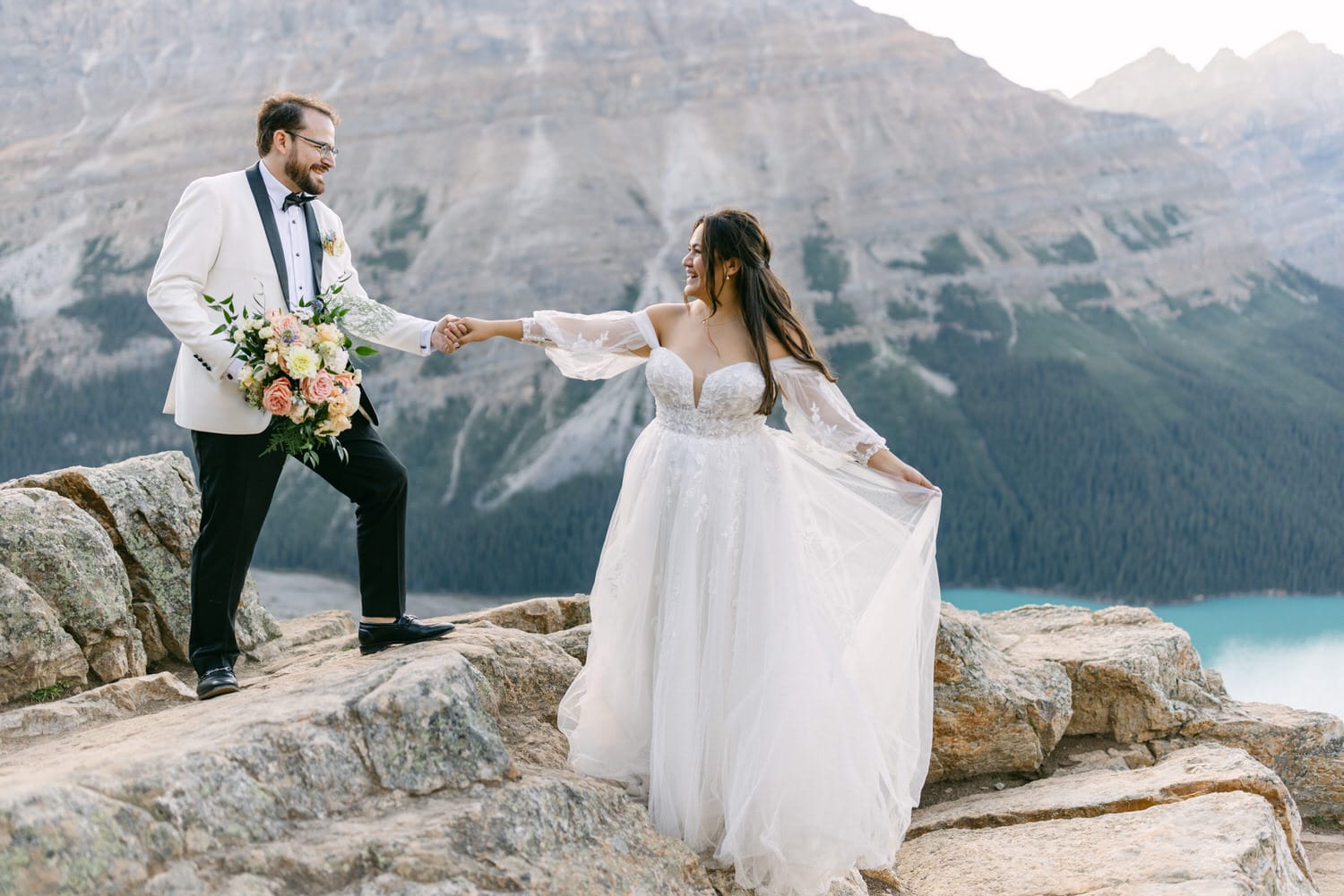 A bride and groom holding hands on a rocky outcrop with a stunning mountainous lake landscape in the background.