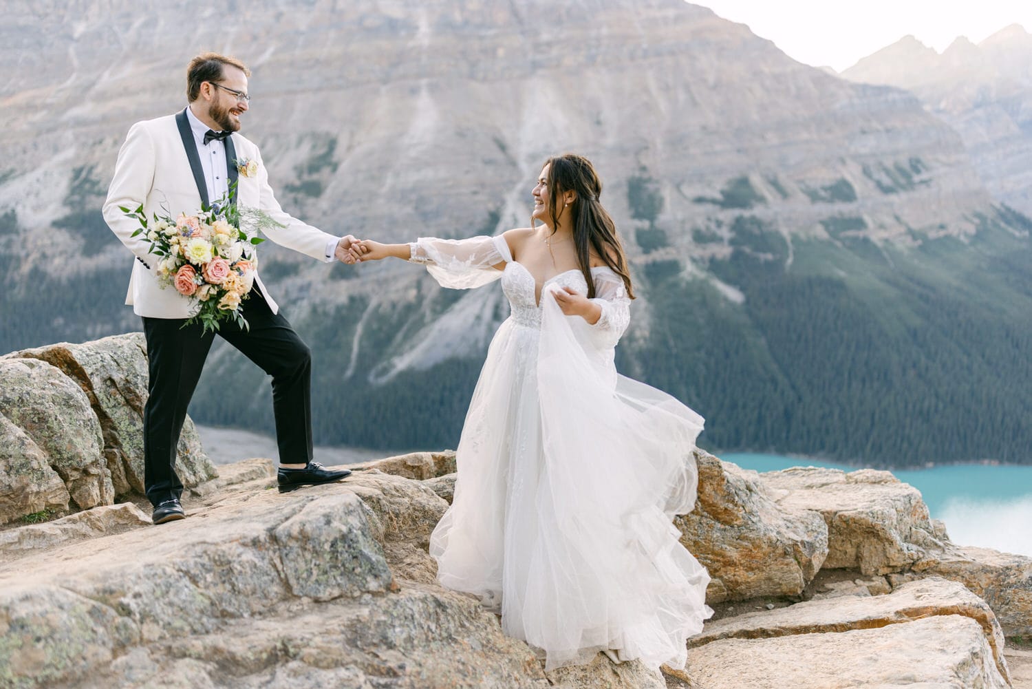 A bride and groom holding hands on a rocky mountain ledge with a serene lake and majestic mountains in the background.