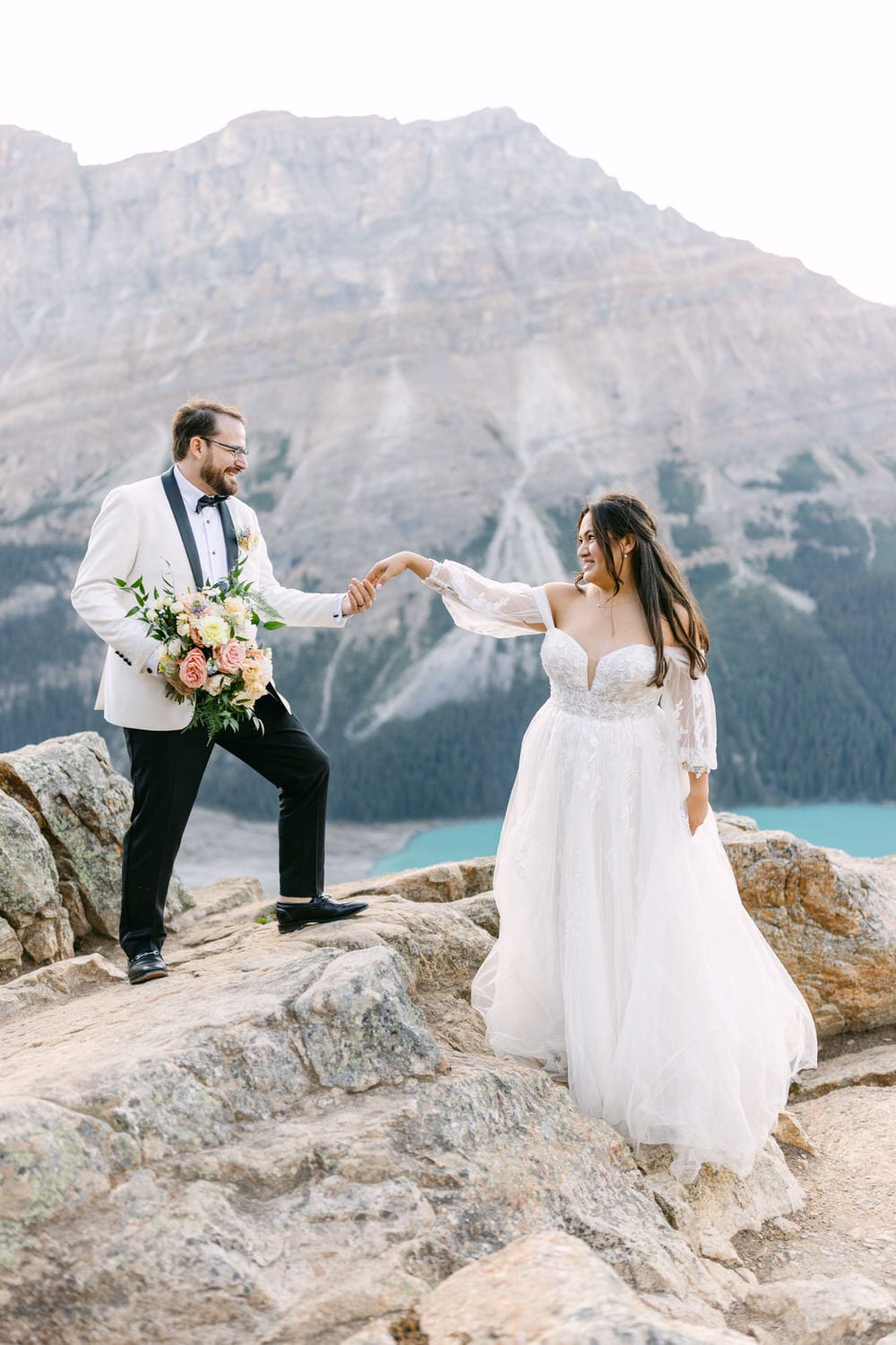 A bride and groom holding hands on rocky terrain with a mountain and turquoise lake in the background.