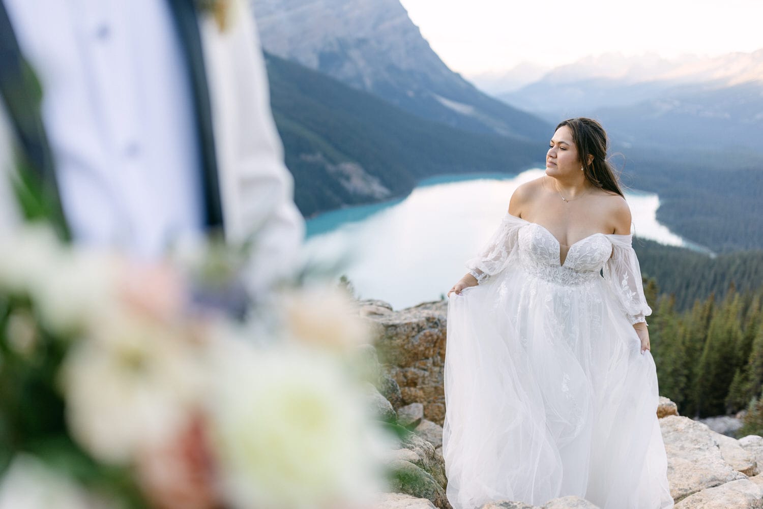 A bride in a white wedding dress with an off-the-shoulder neckline stands in focus against a backdrop of mountains and a turquoise lake, with an out-of-focus groom in the foreground.