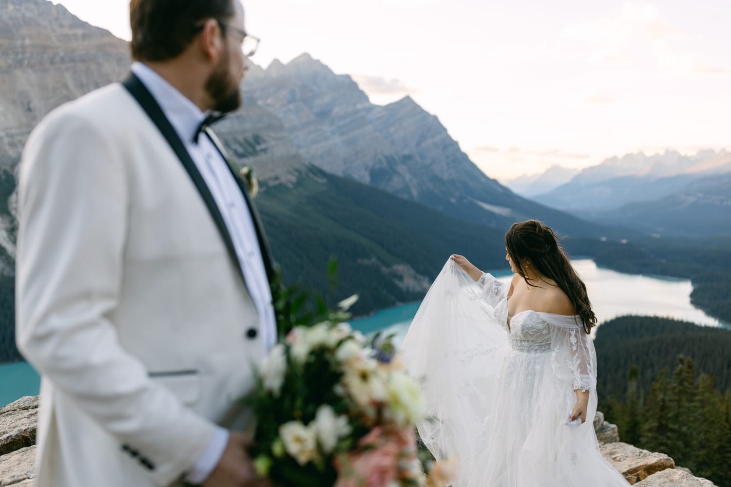 A bride and groom at a scenic mountain overlook, with the bride facing away and the groom partially in the frame, with a beautiful lake and mountains in the background.