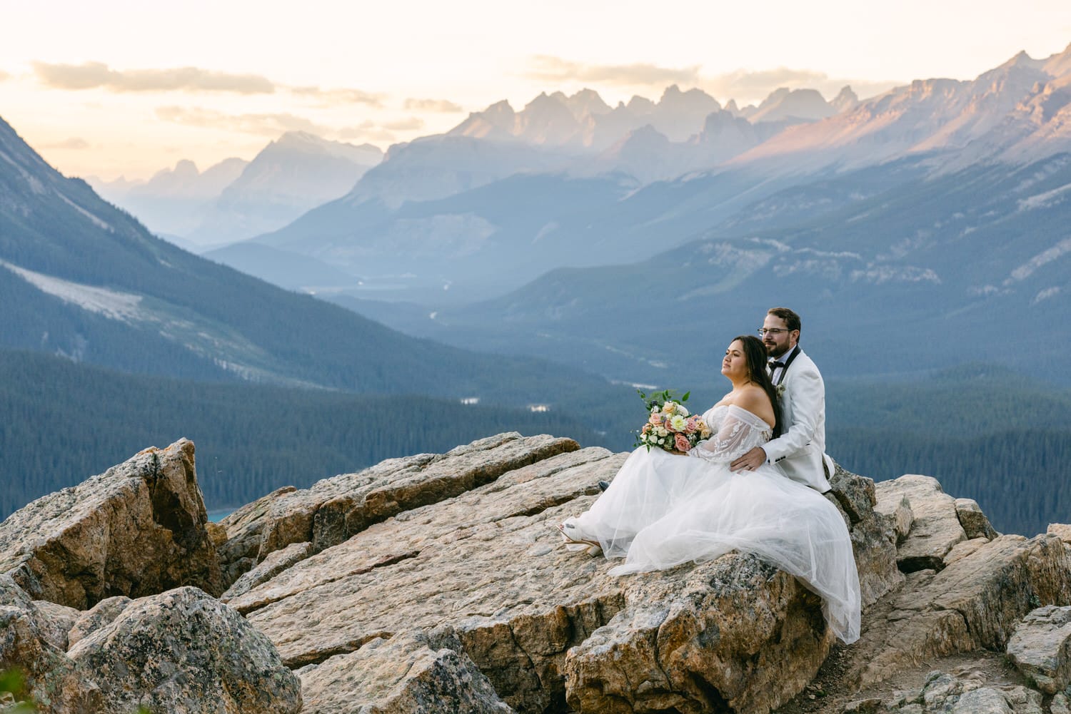 A couple in wedding attire seated on a rocky mountain summit with a panoramic view of distant mountain peaks at sunset.