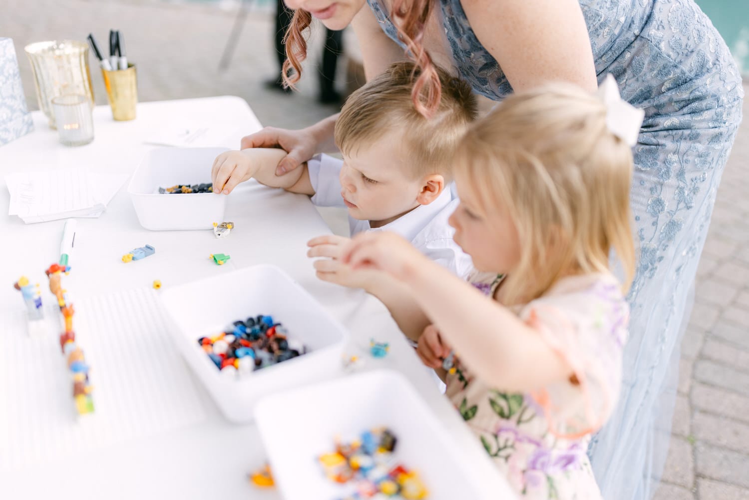 A woman and two children engaged in play with building blocks at a white table outdoors.