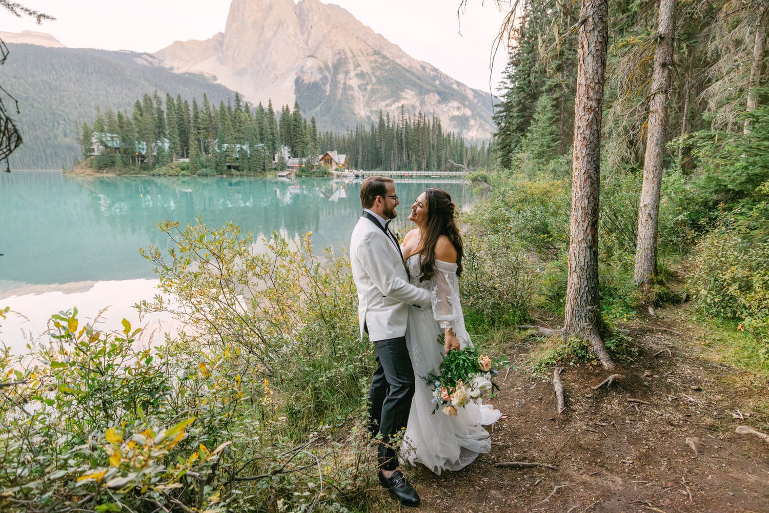 A bride and groom holding hands by a tranquil mountain lake surrounded by forests and mountains.