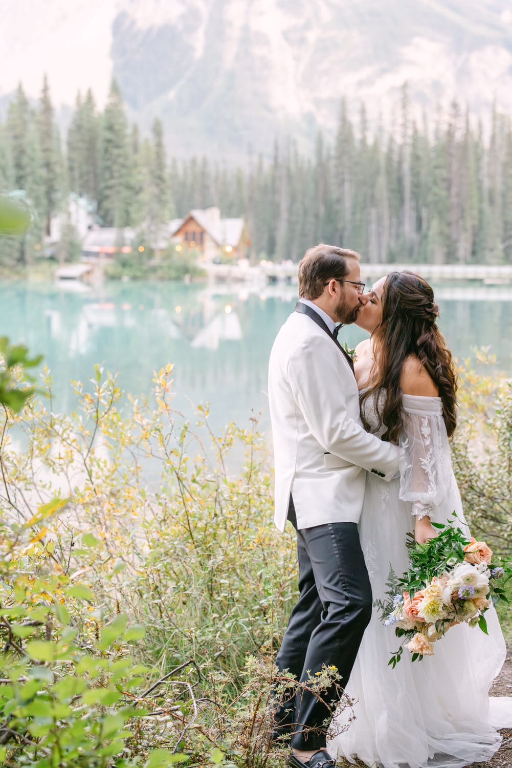 A bride and groom kissing beside an alpine lake with a mountain backdrop and a cabin in the distance.