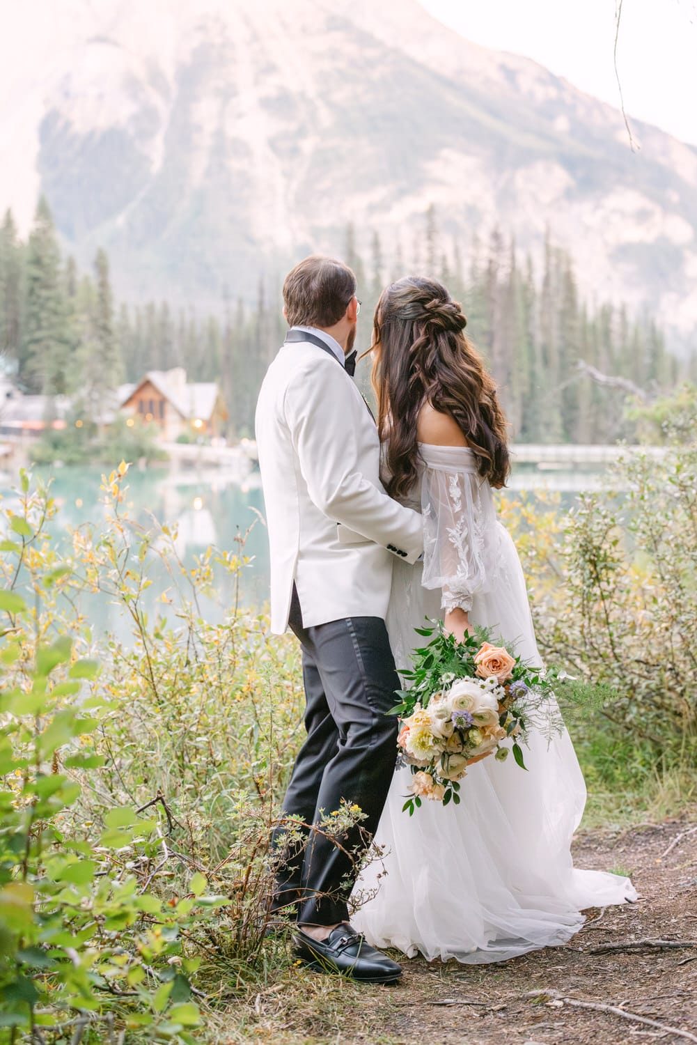 A bride and groom holding each other, facing a serene mountain lake, with the bride holding a bouquet.