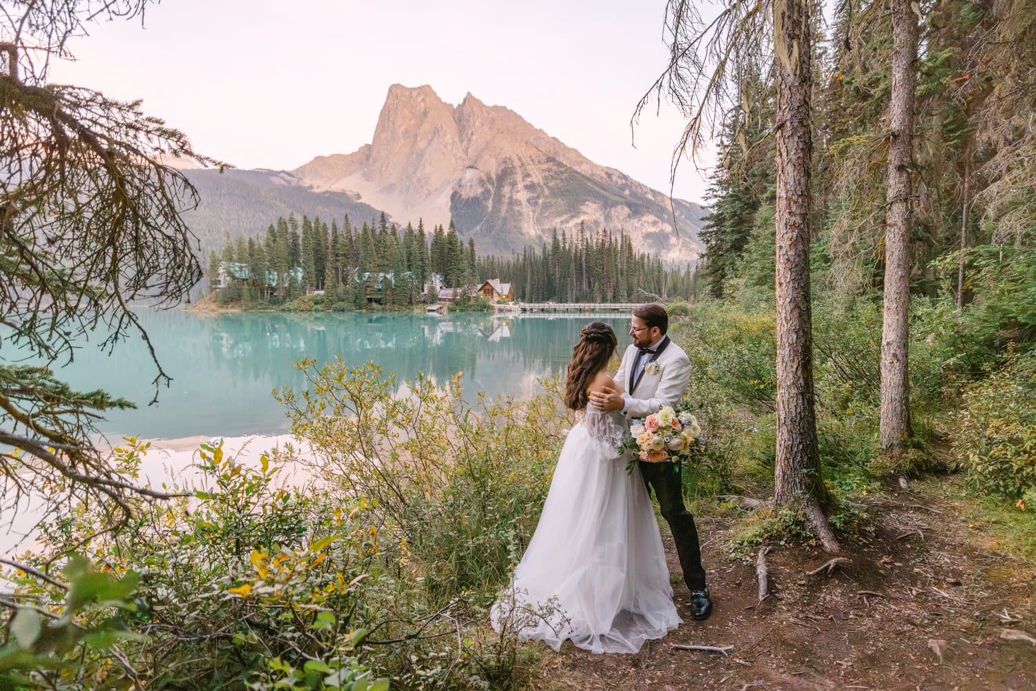 A couple in wedding attire sharing an embrace by a serene mountain lake with forest and peaks in the background.