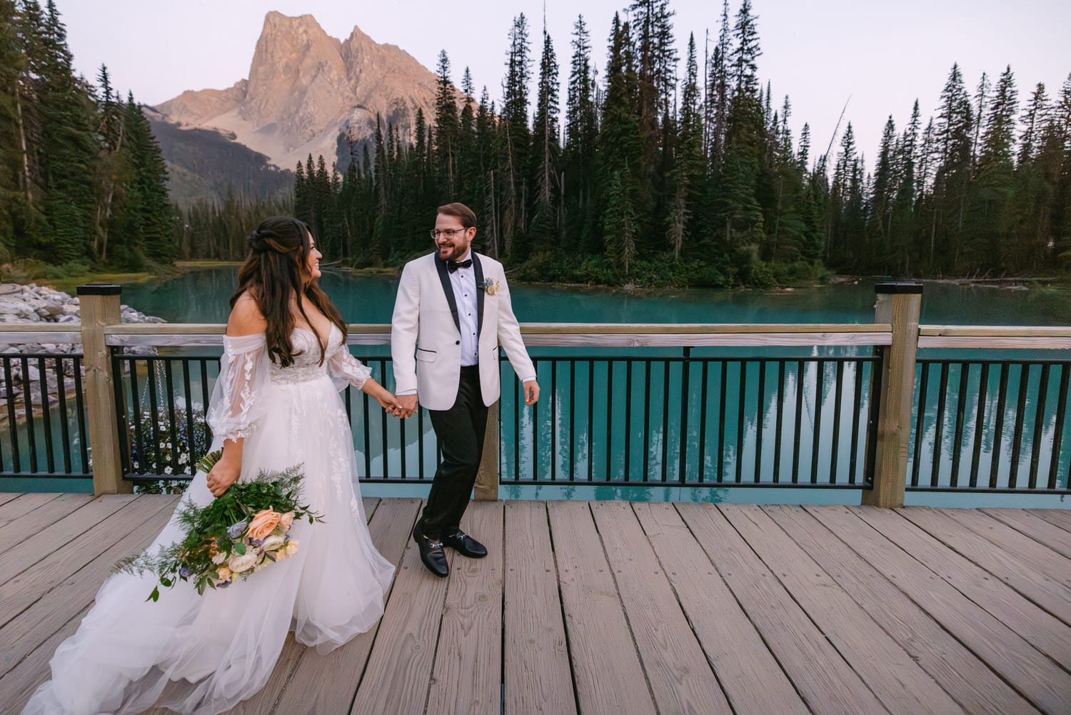 ::A bride and groom holding hands on a wooden deck with a serene turquoise lake and pine trees in the background, mountains looming in the distance during dusk.