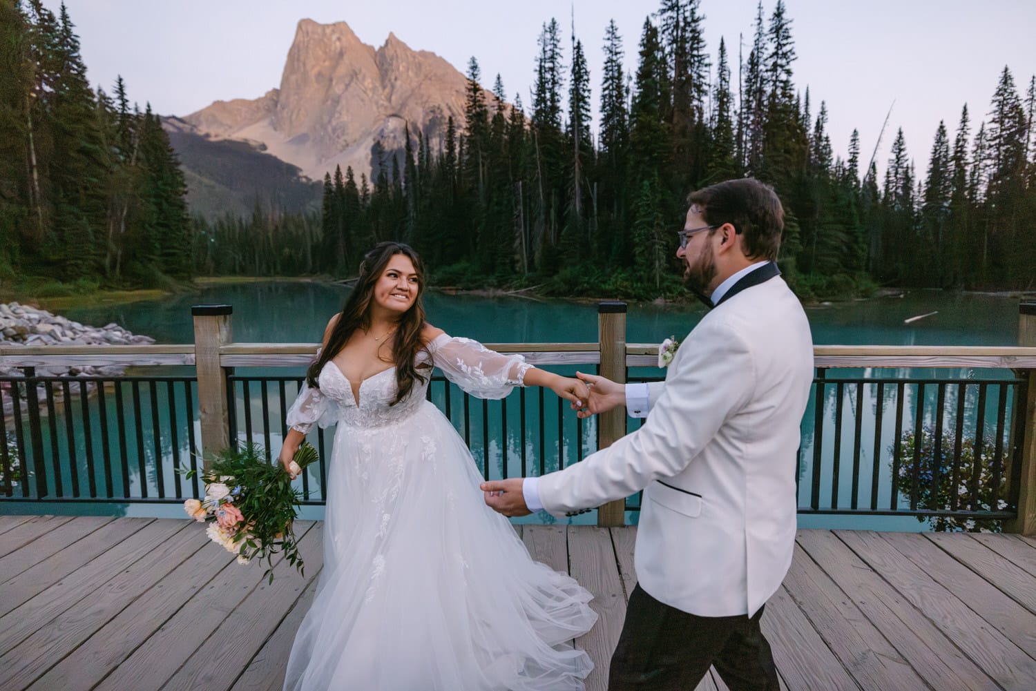 A bride and groom holding hands on a wooden bridge by an alpine lake with a mountain backdrop at dusk.