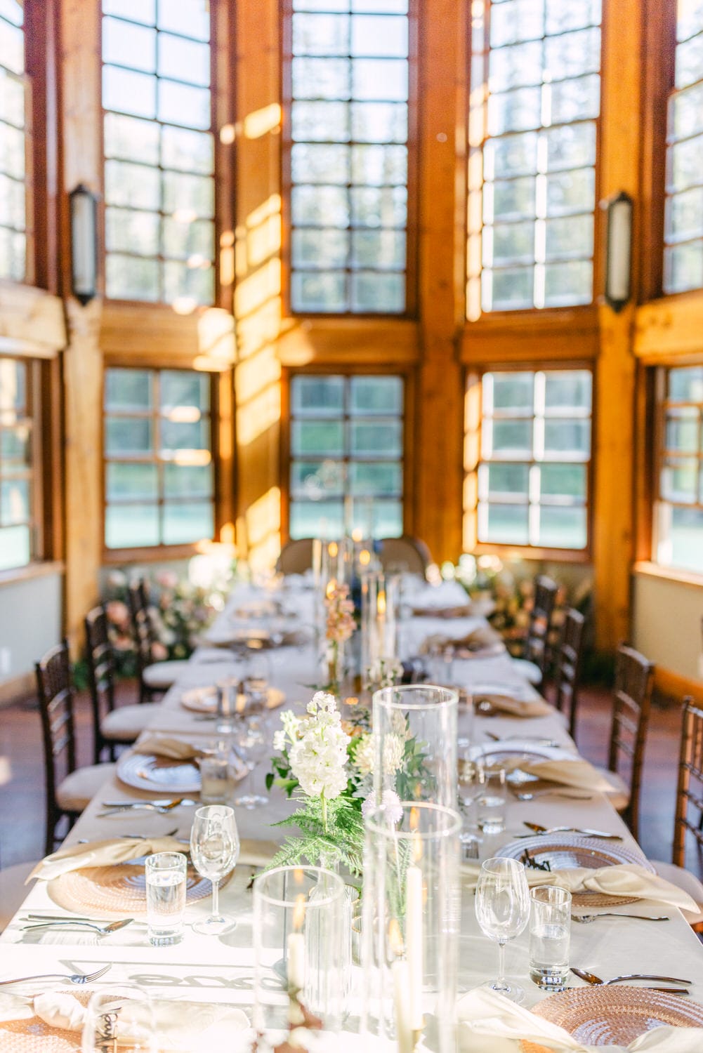 A long dining table set with white linens, crystal glassware, and floral centerpieces in a sunny room with tall windows