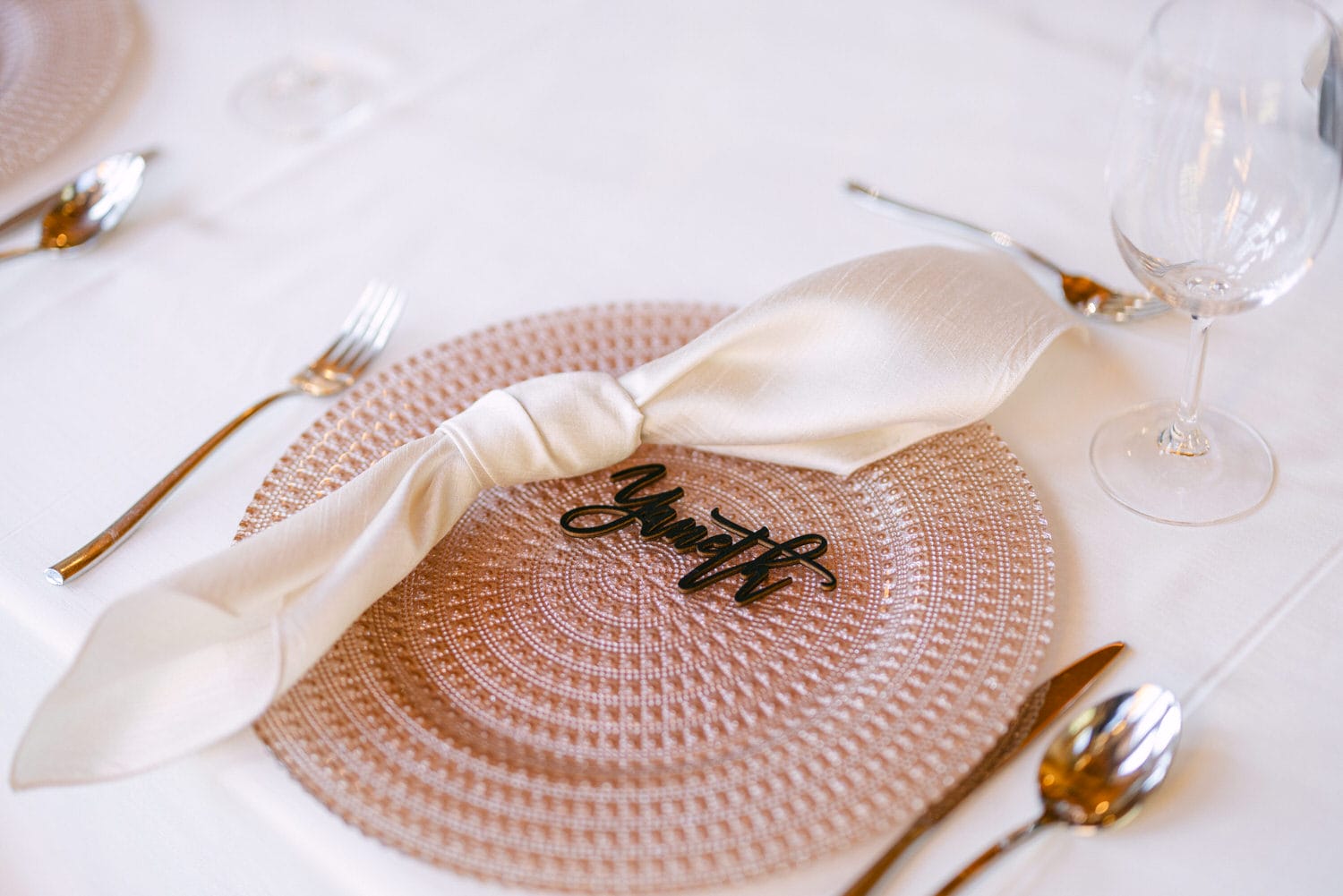 An elegant dining table setting featuring a round beige placemat, a cream napkin with a custom black script name place card, and gold flatware with a clear wine glass on a white tablecloth.