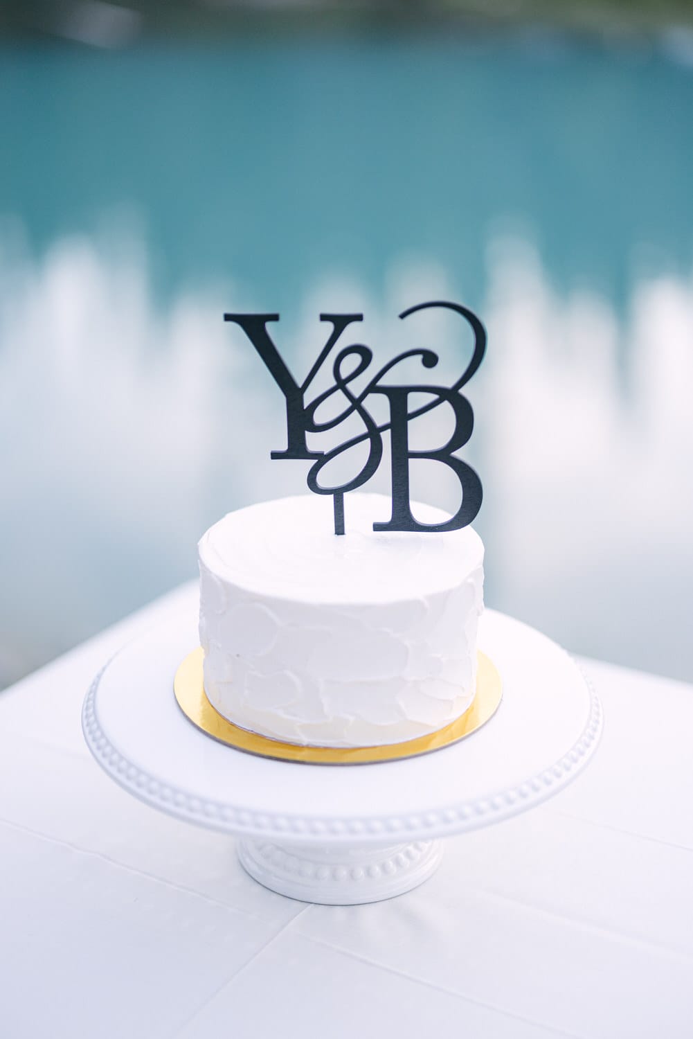 A white cake with a black monogram topper on a round plate with a blurred background.