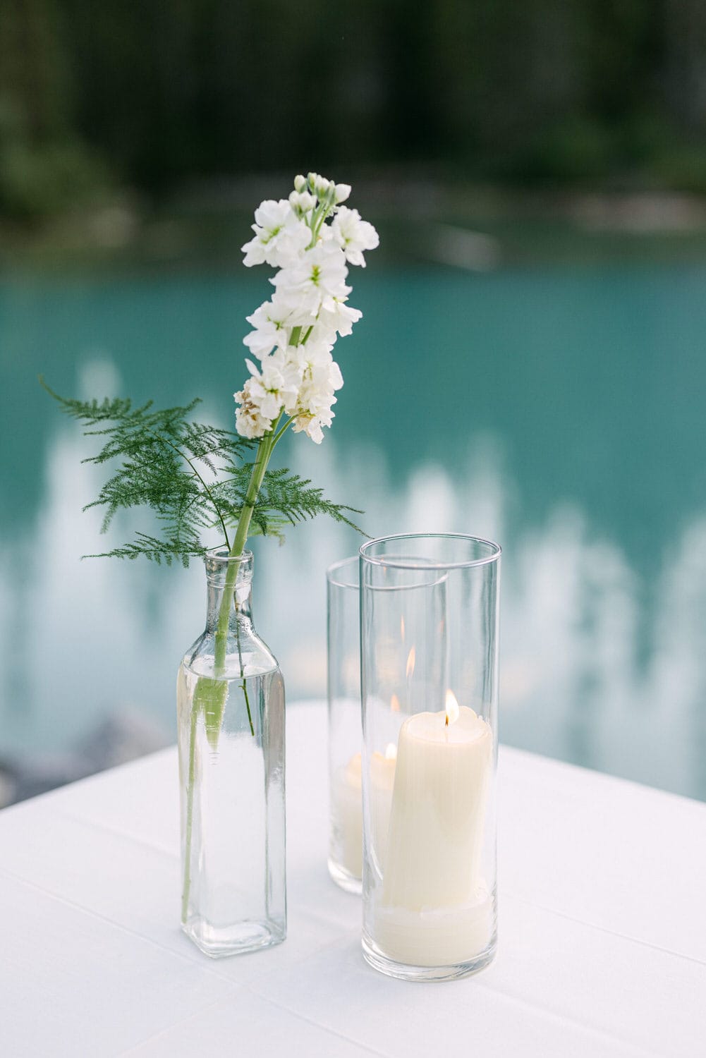 A tranquil setting with a white flower in a clear vase beside two cylindrical glass candle holders with lit candles, all on a table with a serene lake in the background.