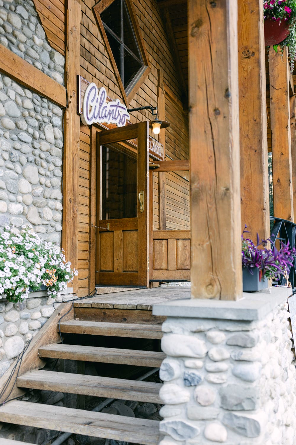 Wooden doorway with a sign reading 'Cilantro,' stone and wood walls, hanging flowers, and steps leading to the entrance.