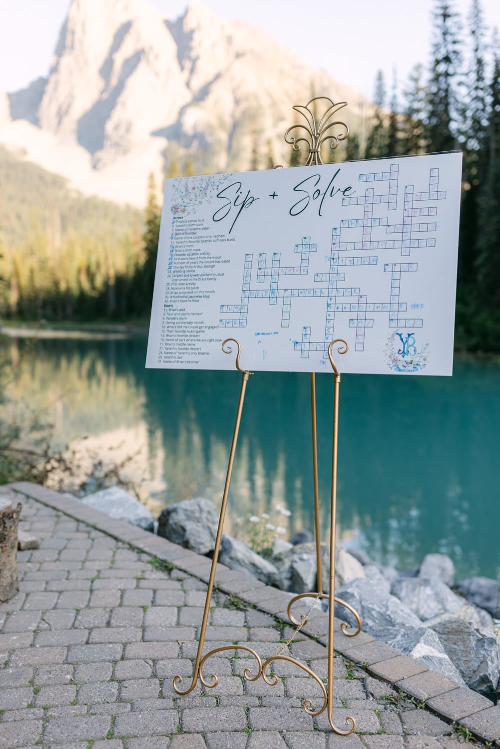 A crossword puzzle sign titled "Sip + Solve" on an elegant stand by a tranquil mountain lake with pine trees and a mountain peak in the background.