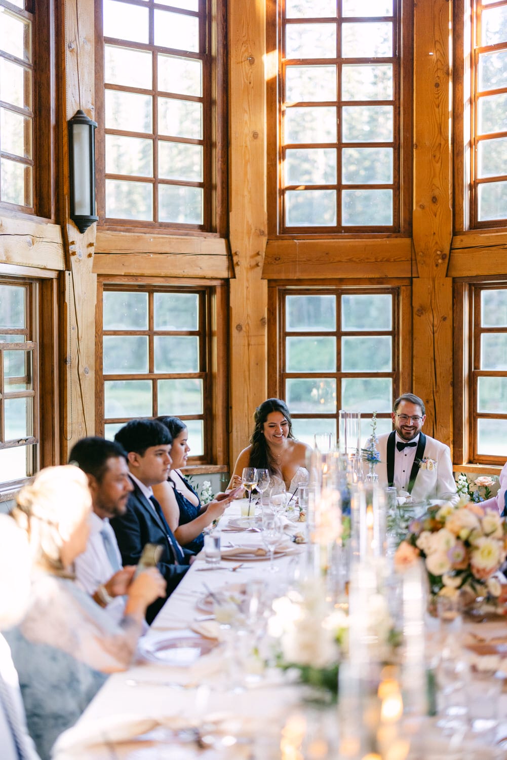 Guests enjoying a meal at a wedding reception inside a room with wooden walls and windows showing trees outside, table set with flowers and candles.