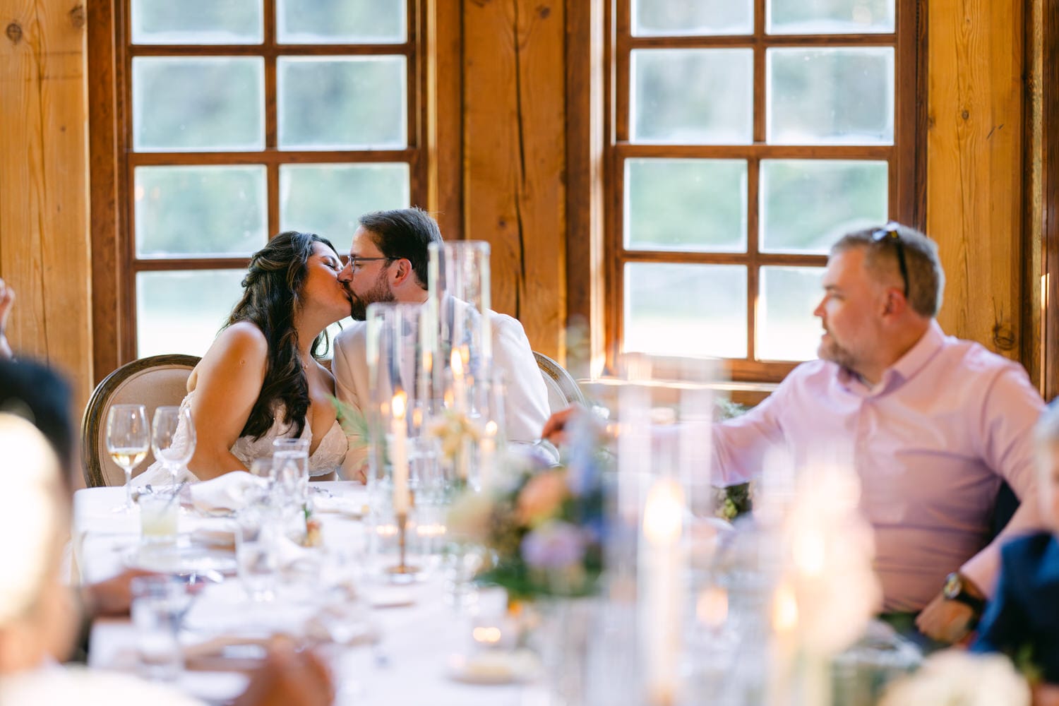 A bride and groom sharing a kiss at their wedding reception table, with guests seated around them and elegant table settings in view.