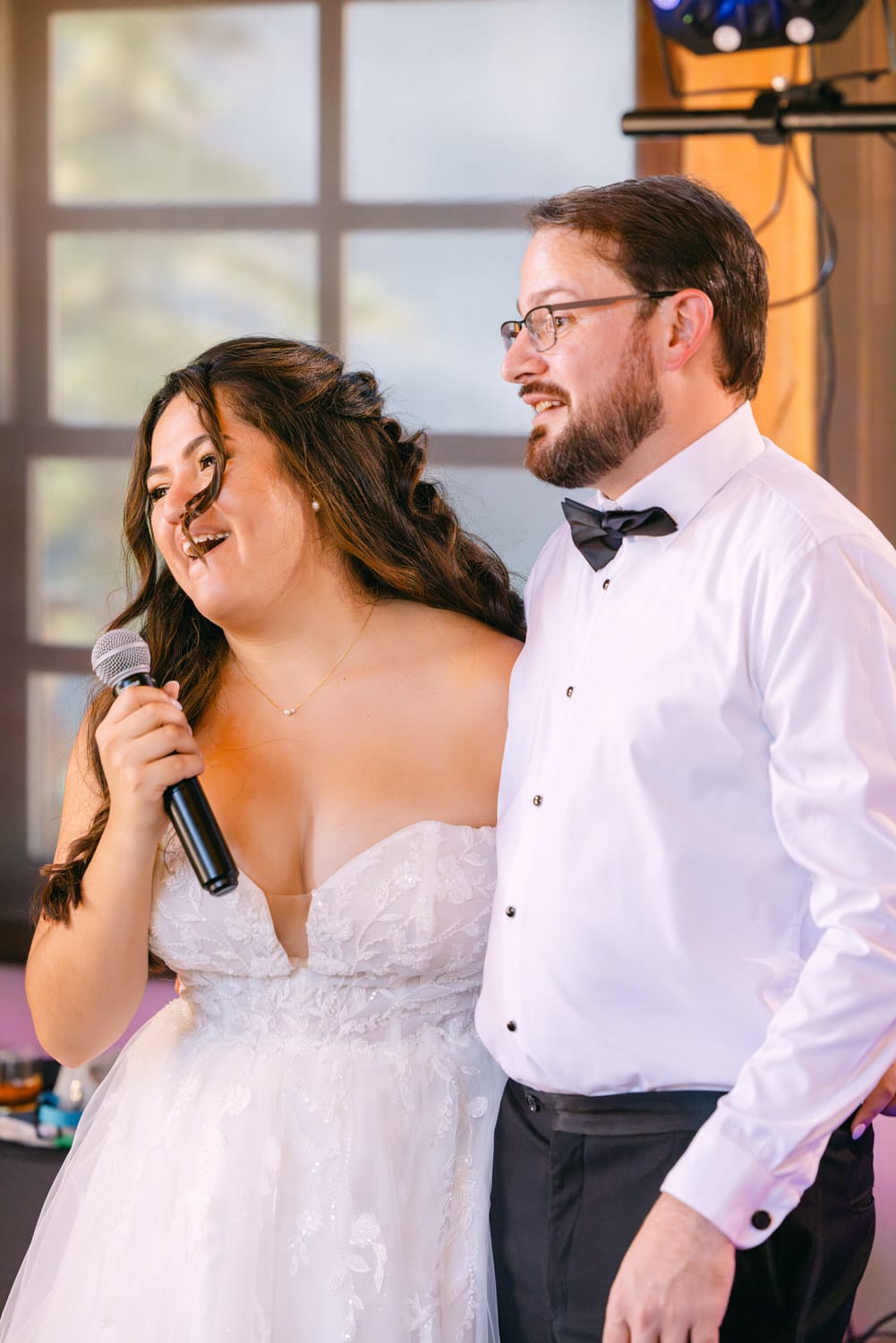 A bride holding a microphone and smiling mid-speech beside a man in formal attire during a wedding reception.