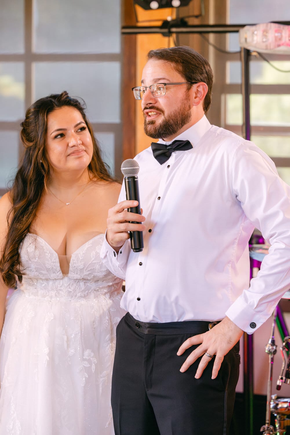 Man in a white shirt and bow tie speaking into a microphone with a woman in a white wedding dress standing beside him.