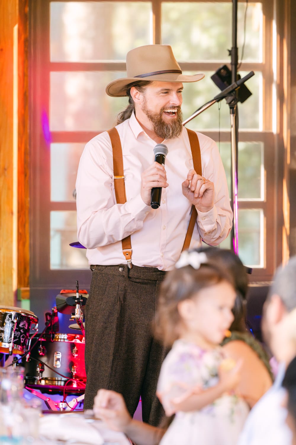 A smiling bearded man wearing a hat and suspenders holding a microphone and speaking at a social gathering inside a room with red-tinted windows.