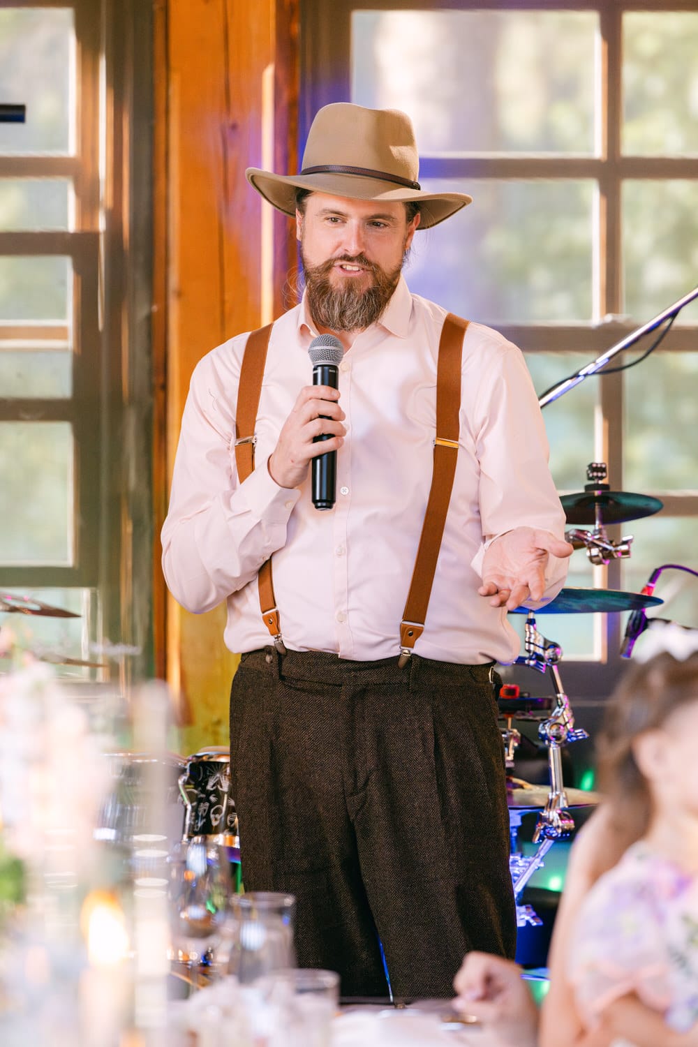 A man with a beard wearing a hat, suspenders, and holding a microphone appears to be giving a speech in a warmly lit room.