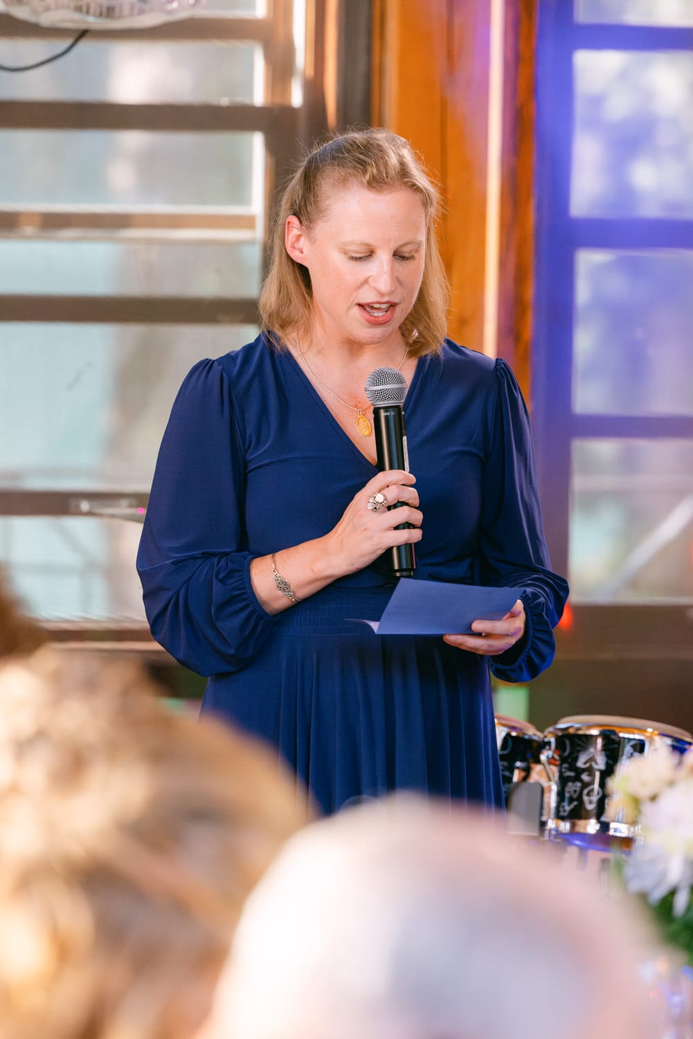 A woman in a blue dress speaking into a microphone while reading from a note card, with an audience in the foreground.